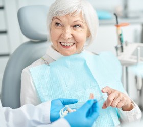 Gloved hands handing replacement teeth to woman with white hair in dental chair