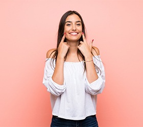 Woman pointing to her dental crowns in Bloomfield with pink background.